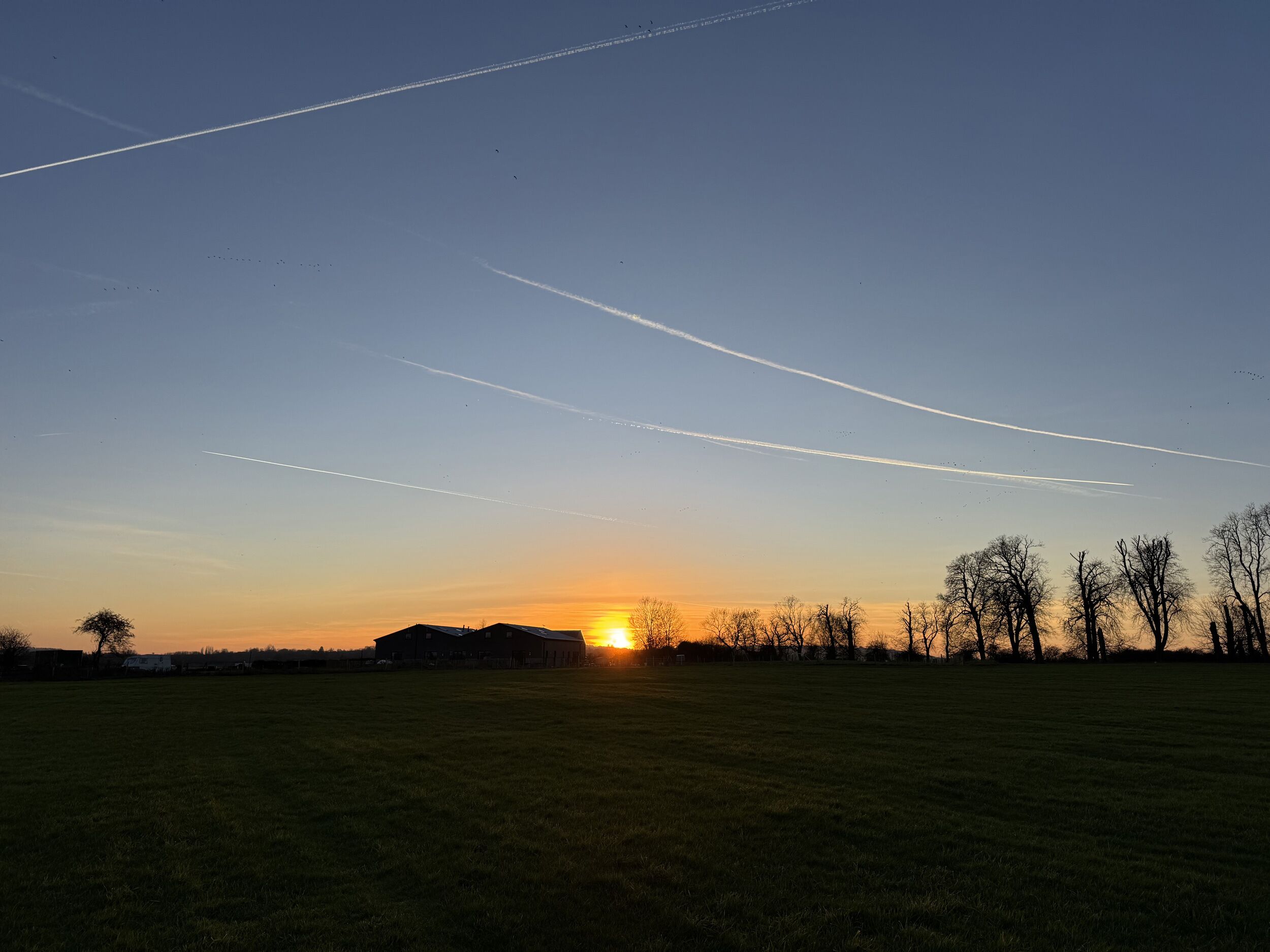 Un atardecer sobre un campo cubierto de hierba, con un edificio a lo lejos y estelas en el cielo.