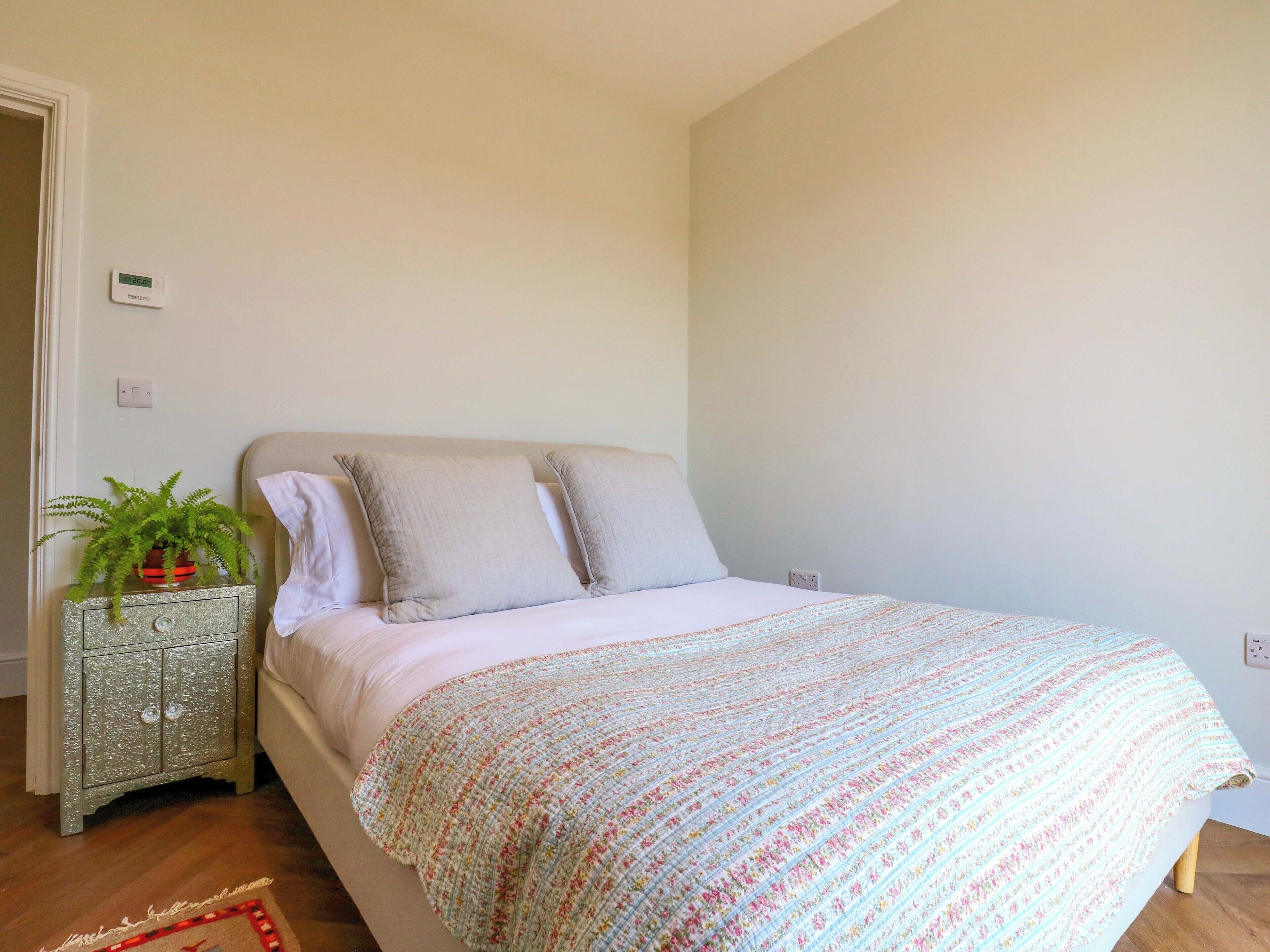 A bedroom with a bed, a nightstand with a potted plant, and a wall thermostat.