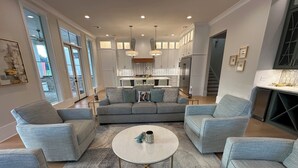 A modern living room with a grey sofa set, a round coffee table, and a kitchen in the background. - The Kentwood- A Luxury Home in Baton Rouge- minutes from LSU and downtown! (Baton Rouge)