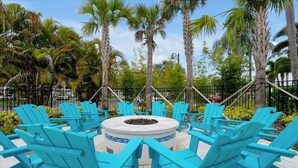 Outdoor seating with chairs arranged around a central fire pit, surrounded by palm trees and a fence.