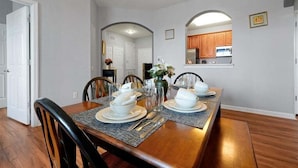 A dining area with a wooden table set for four, surrounded by chairs. There is a kitchen visible through an archway in the background.