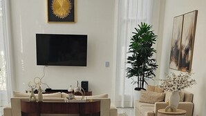 Een eetgedeelte met een grote tafel gedekt voor het ontbijt, een aan de muur bevestigde televisie, en een woonkamer met een bank en een plant.