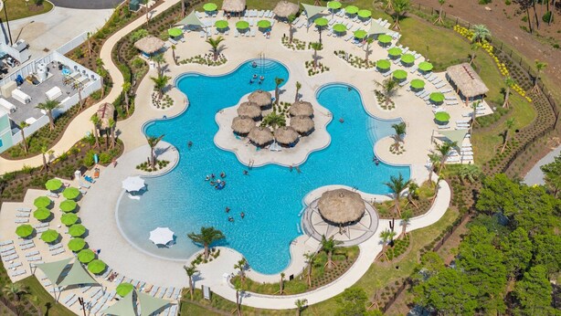 Vue aérienne d’un complexe hôtelier doté d’une piscine centrale, entourée de transats et de parasols, ainsi que d’un espace paysager agrémenté de palmiers.