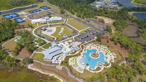 Vue aérienne d’un complexe hôtelier comprenant un bâtiment central, une piscine, des courts de tennis et un parking.