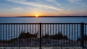 Een zonsondergang vanaf een balkon met een hekwerk, met uitzicht op een wateroppervlak en een land in de verte.