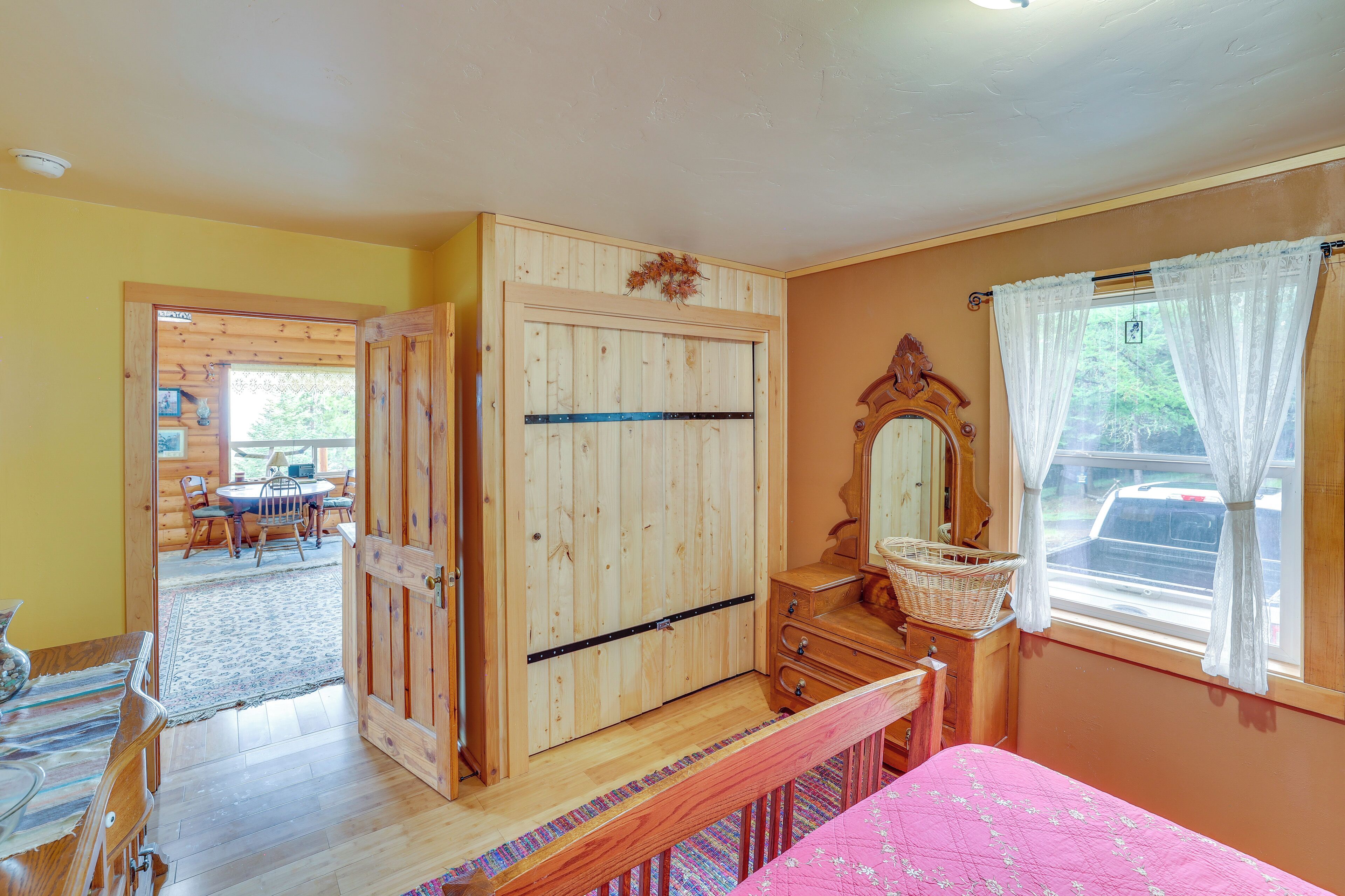 Een kamer met een houten garderobe, een bed met een roze beddeken, een houten dressoir, een rieten mand en een raam met gordijnen.