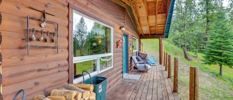 A wooden cabin with a porch, log storage, and a view of the forest.