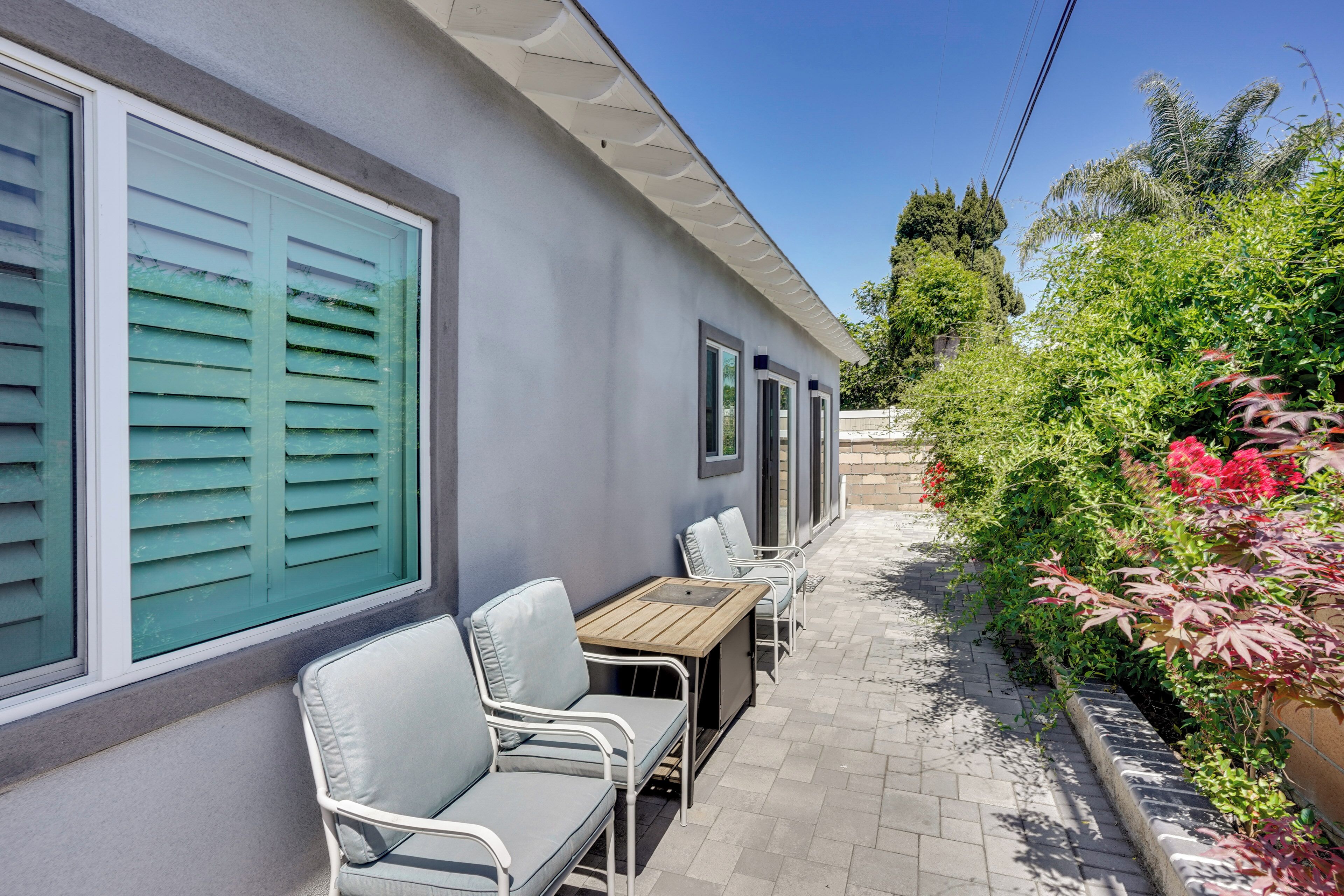 A patio with a table and chairs, a window with shutters, and a garden with plants and flowers.