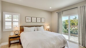 A bedroom with a large bed, a nightstand, a bedside table, a window with blinds, and a view of greenery outside. - 2 Mi to Disneyland: Home w/ Furnished Patio! (Anaheim)