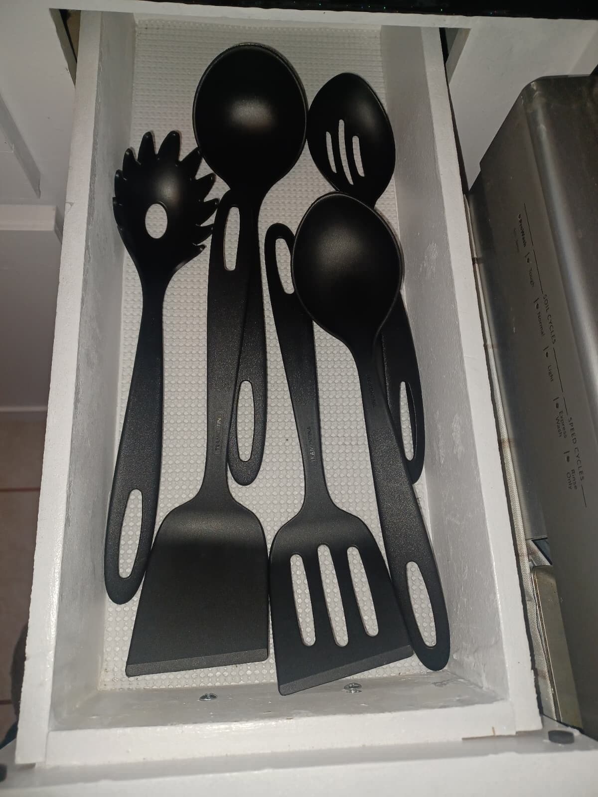 Un cassetto pieno di utensili da cucina neri, tra cui una spatola, un cucchiaio e una forchetta.