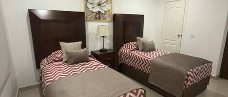 A hotel room with two beds, a nightstand, a lamp, and a door.