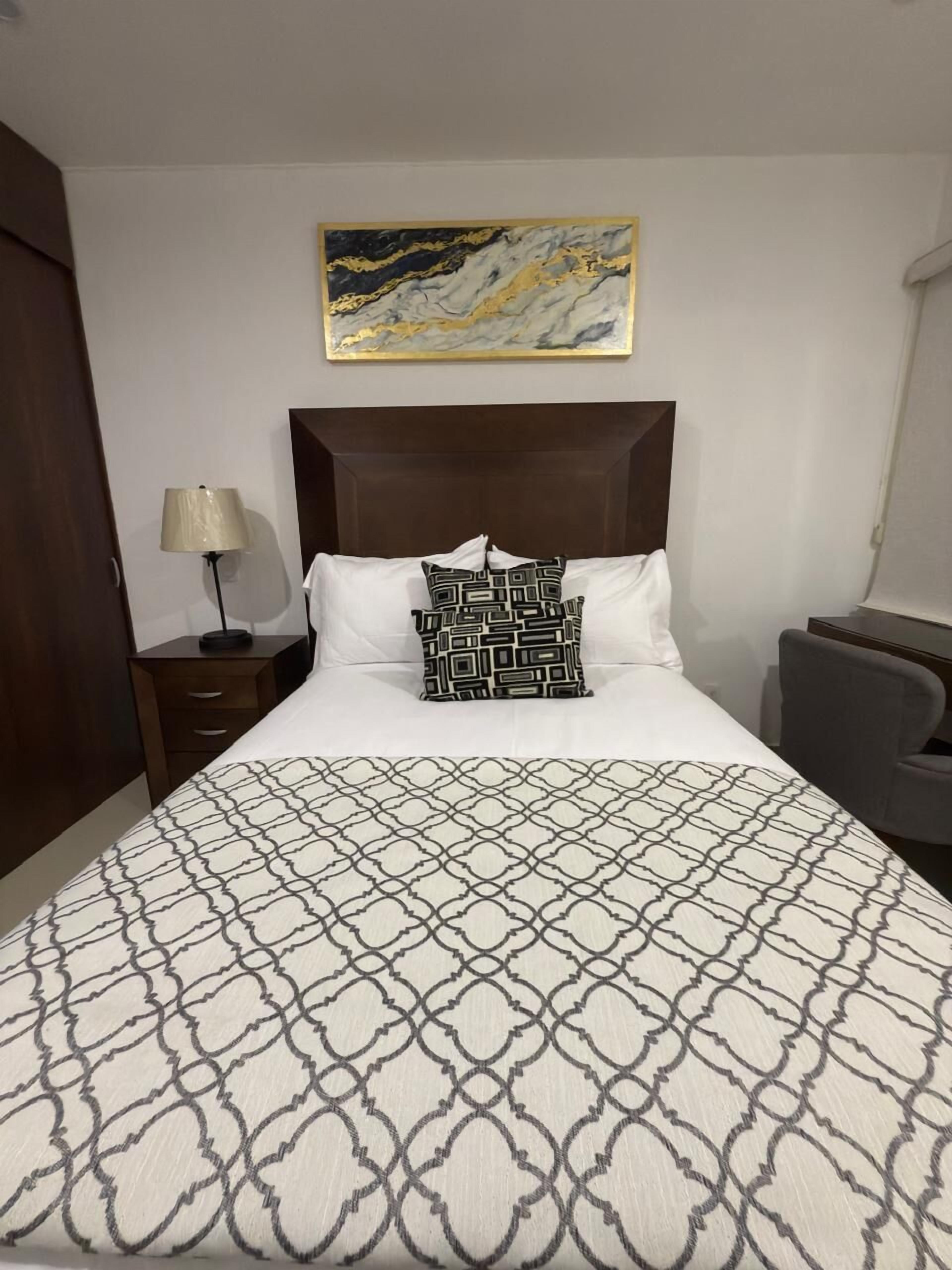 A hotel room with a large bed, a headboard, a nightstand, a lamp, a chair, and a painting on the wall.