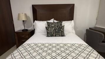 A hotel room with a large bed, a headboard, a nightstand, a lamp, a chair, and a painting on the wall.
