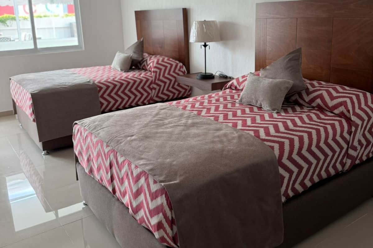 Habitación de hotel con dos camas, cada una con cabecera de madera y colcha estampada. Ventana que ofrece luz natural y vistas al exterior.