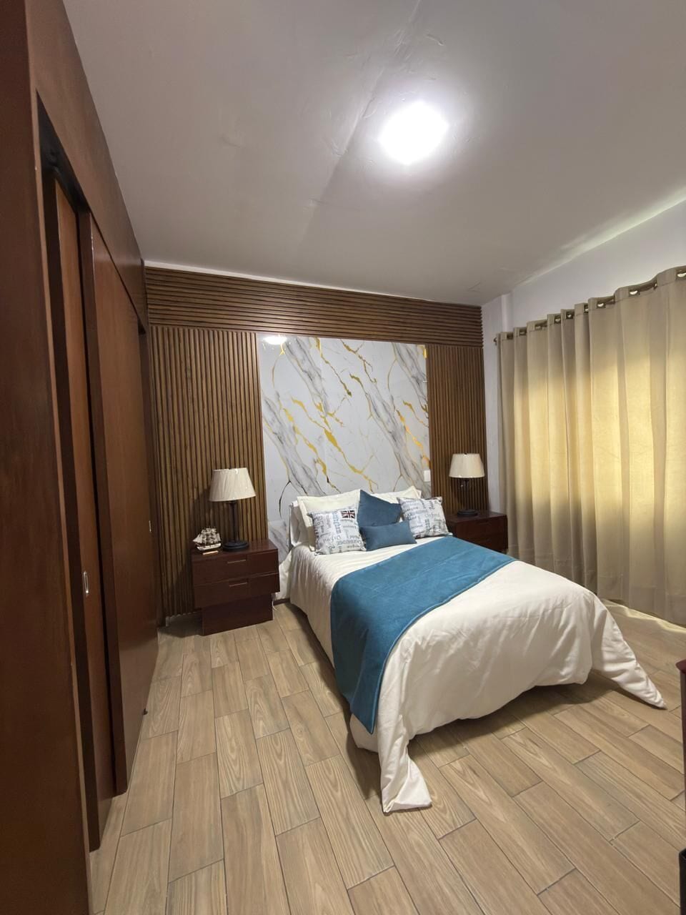 A hotel room with a bed, bedside tables, a lamp, curtains, and a wooden floor.