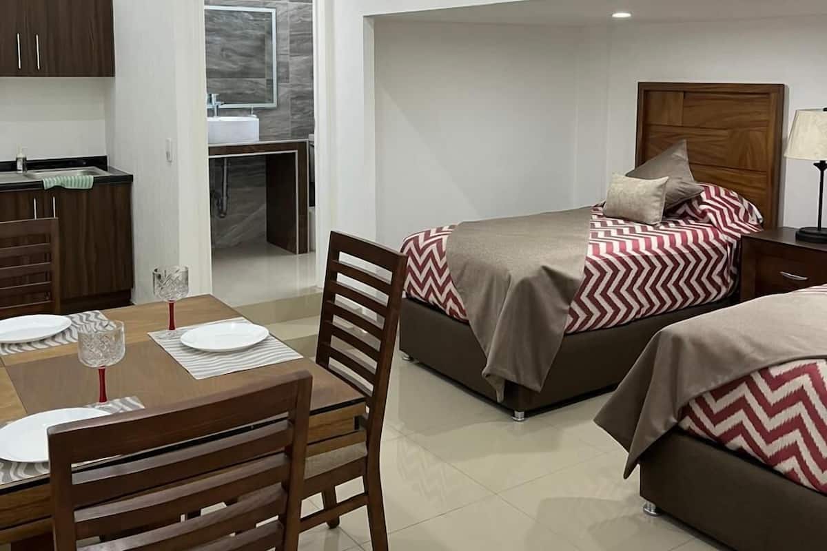 Una habitación de hotel moderna y compacta con comedor, dos camas con ropa de cama estampada, una kitchenette pequeña y un baño con espejo y lavamanos.