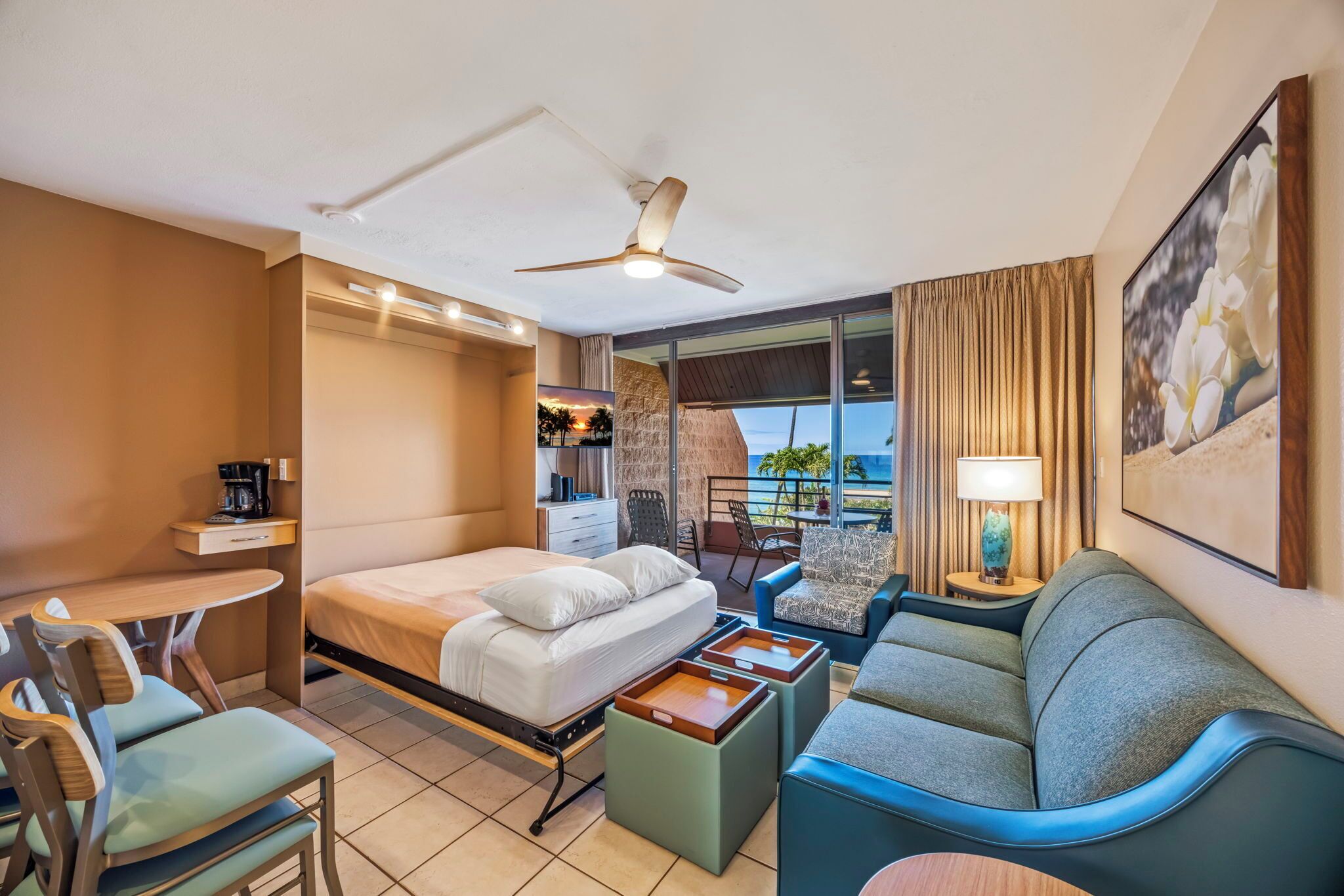 A hotel room with a bed, a desk, a sofa, a balcony with a view, and a ceiling fan.