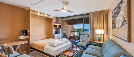 A hotel room with a bed, a desk, a sofa, a balcony with a view, and a ceiling fan.