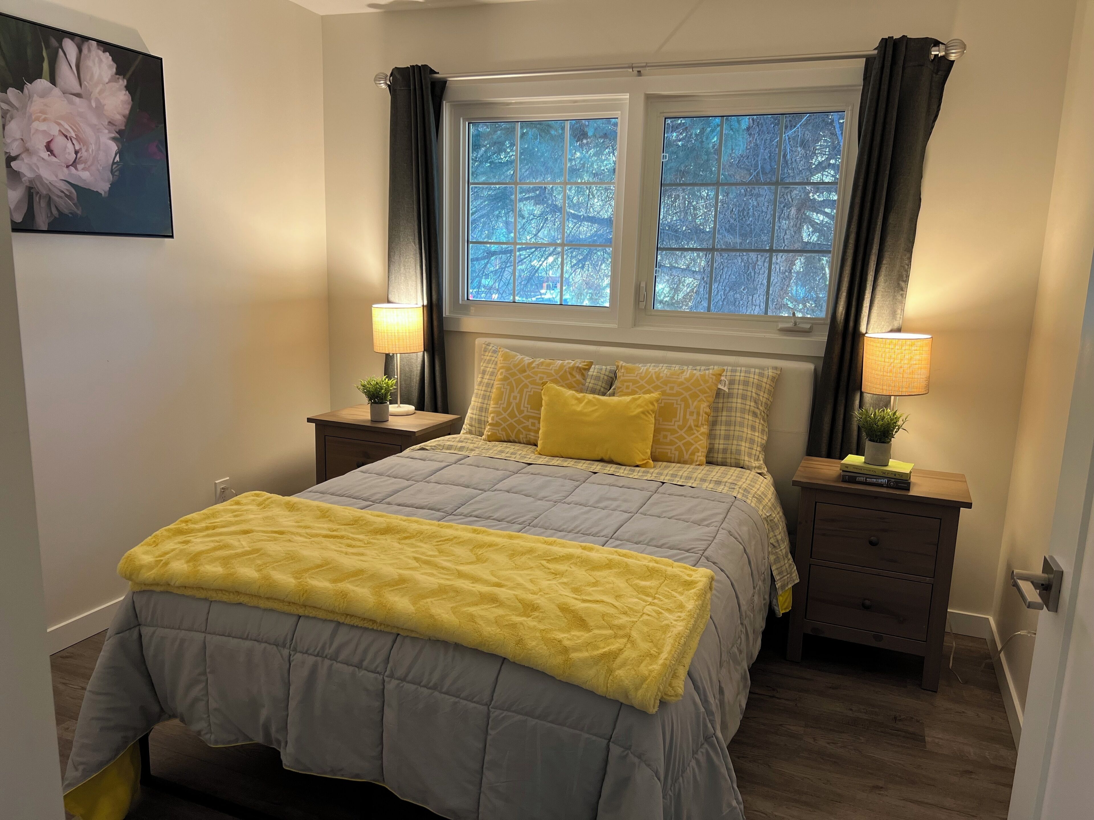 Une chambre à coucher avec un lit, deux tables de chevet, une fenêtre avec des rideaux et un tableau au mur.