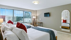 Ein Hotelzimmer mit einem großen Bett, einem Fernseher, einem Schreibtisch und einem Fenster mit Blick auf die Stadt.