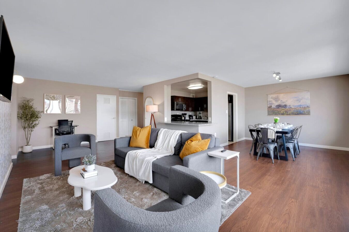 A modern living room with a grey sofa, a dining table with chairs, a small white table, and a kitchen area in the background.