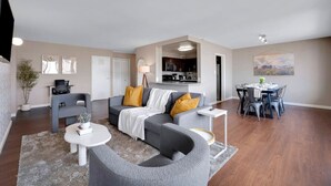 Ein modernes Wohnzimmer mit einem grauen Sofa, einem Esstisch mit Stühlen, einem kleinen weißen Tisch und einer Küche im Hintergrund.