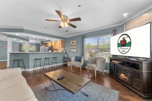 A modern living room with a fireplace, a ceiling fan, and a kitchen in the background. - Relaxing Walk-In Condo •Community Pool • Pet Friendly (Branson)
