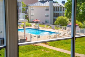 Une piscine avec des chaises longues et un parasol rouge, entourée d’une zone délimitée par une clôture et d’un bâtiment doté de nombreuses fenêtres.