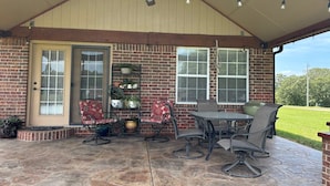A covered patio with a brick wall, a door, and a window. There are four chairs and a table with a chair.