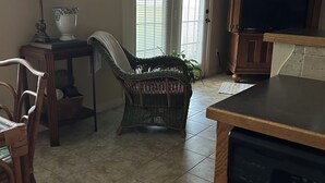A dining area with a table, chairs, a wicker chair, a lamp, and a television in the background.
