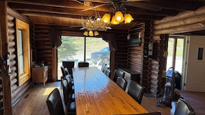 Ein Essbereich in einer Holzblockhütte mit einem langen Holztisch, schwarzen Ledersesseln, einer Chandelierleuchtre und Blick ins Freie.