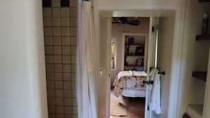 A narrow hallway with a wooden floor leading to a bedroom with a bed and a nightstand. A white curtain is partially drawn, and there is a ceiling fan visible. - Historic Mill House Unit 3 (Taos)