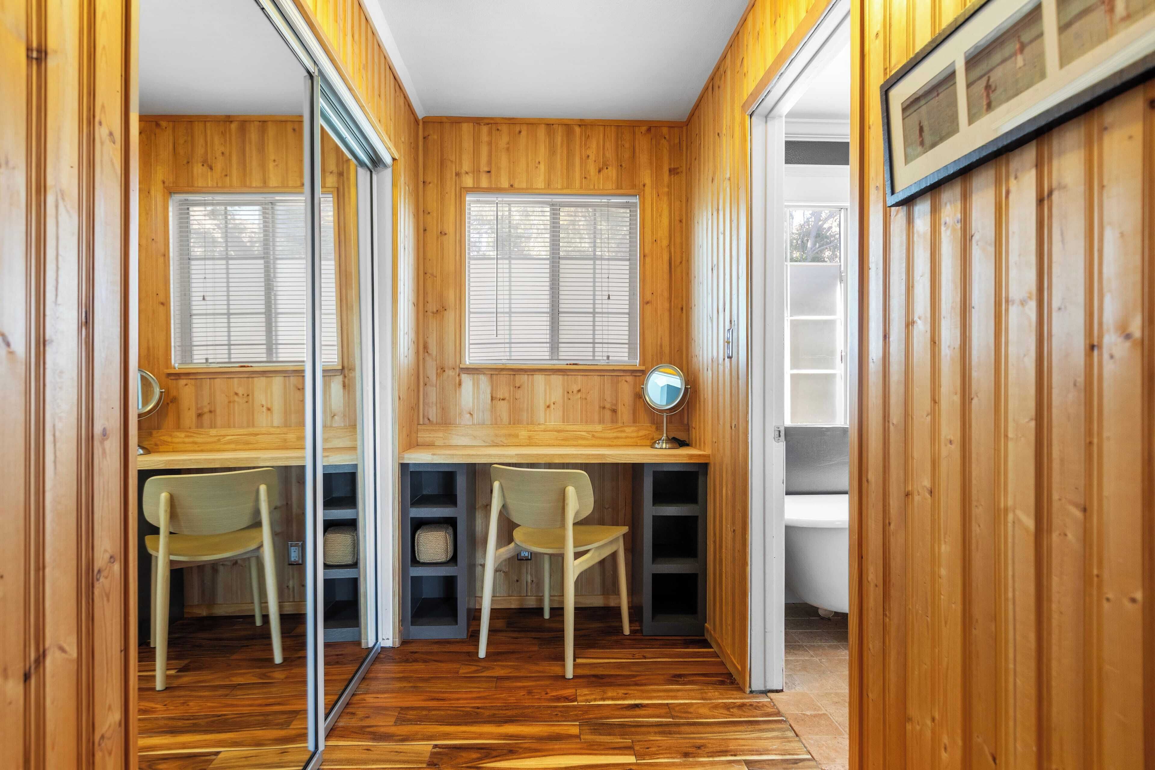 A room with wooden walls and floors, two desks with chairs, a mirror, and a window.