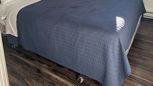 Una cama bien hecha con colcha azul, cabecera de madera y una ventana con cortinas.