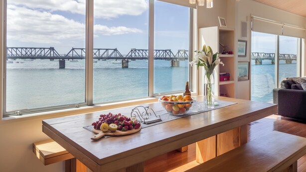 Una cocina moderna con amplios ventanales que dan a un río y un puente, una mesa de madera con fruta y un jarrón de flores.