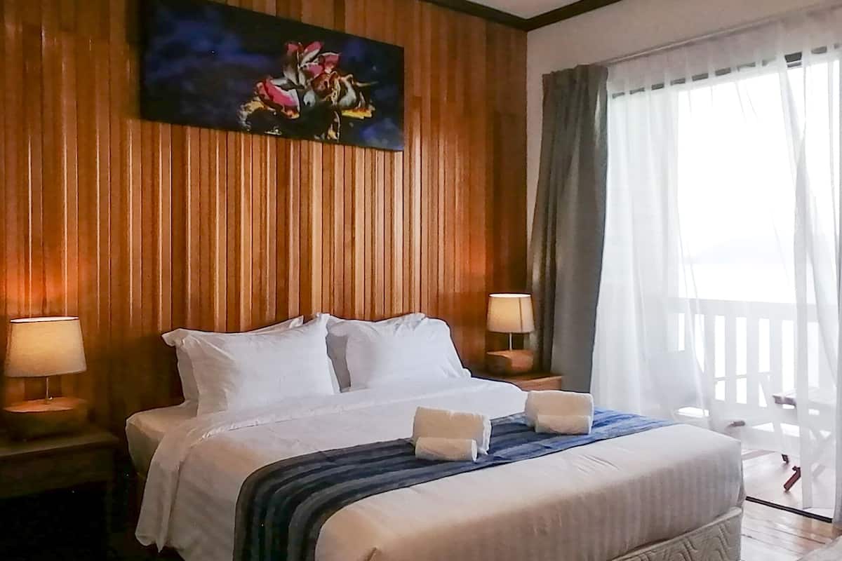 A hotel room with a bed, two bedside lamps, a painting on the wall, and a view of the outdoors through a window with curtains.
