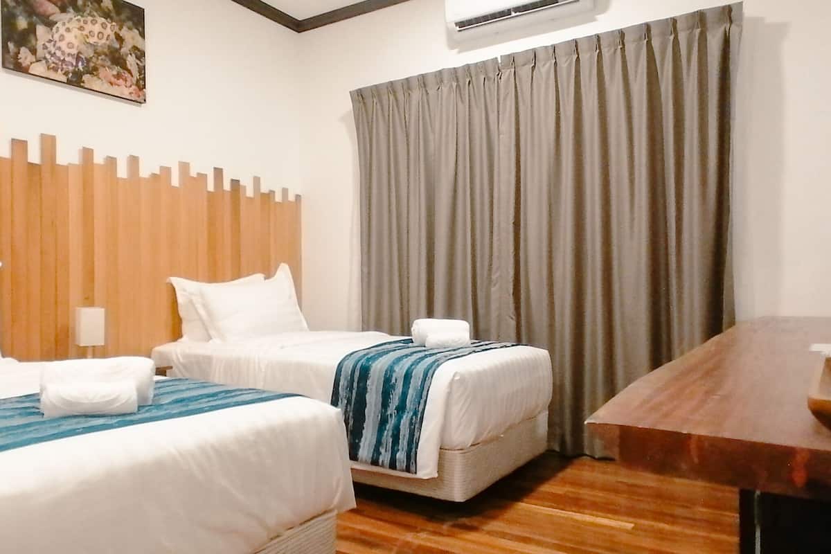 A hotel room with two beds, a wooden headboard, a large window with curtains, a wooden desk, and a wall-mounted air conditioner.