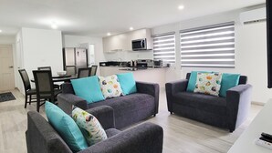 Ein modernes Wohnzimmer mit grauen Sofas, Türkis-Kissen, einem Essbereich mit Tisch und Stühlen sowie einer Küche mit Geräten.