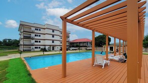 Eine Holzterrasse mit Pool und Liegestühlen, im Hintergrund ein mehrgeschossiges Gebäude und ein klarer Himmel.
