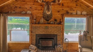 A rustic cabin interior with a large deer head mounted above a fireplace, wooden walls, and a view of a lake through two windows. - The Moose Lodge on the Susquehanna River (Sugar Run)