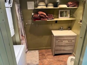 Un baño compacto con tocador de madera, inodoro y repisas sobre el tocador con toallas y artículos de aseo.