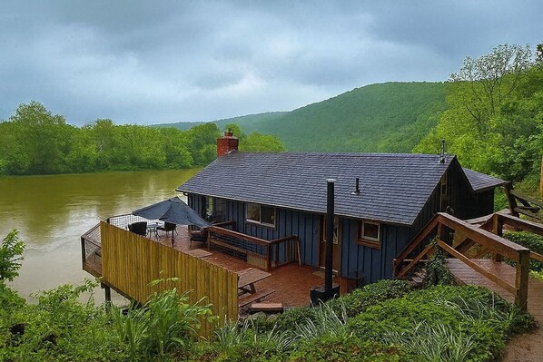 Una cabaña de madera con terraza que da al río, rodeada de vegetación y colinas.