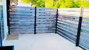 Uma área externa coberta com piso de concreto, grades de metal e vista para um estacionamento e árvores.