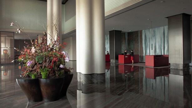 Un hall d’entrée spacieux agrémenté d’un grand ensemble de plantes artificielles, de sols en marbre poli et de comptoirs d’accueil rouges.