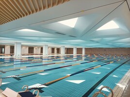 Una gran piscina interior con varias pistas de natación, un trampolín y una zona de estar, techos altos y luz natural.