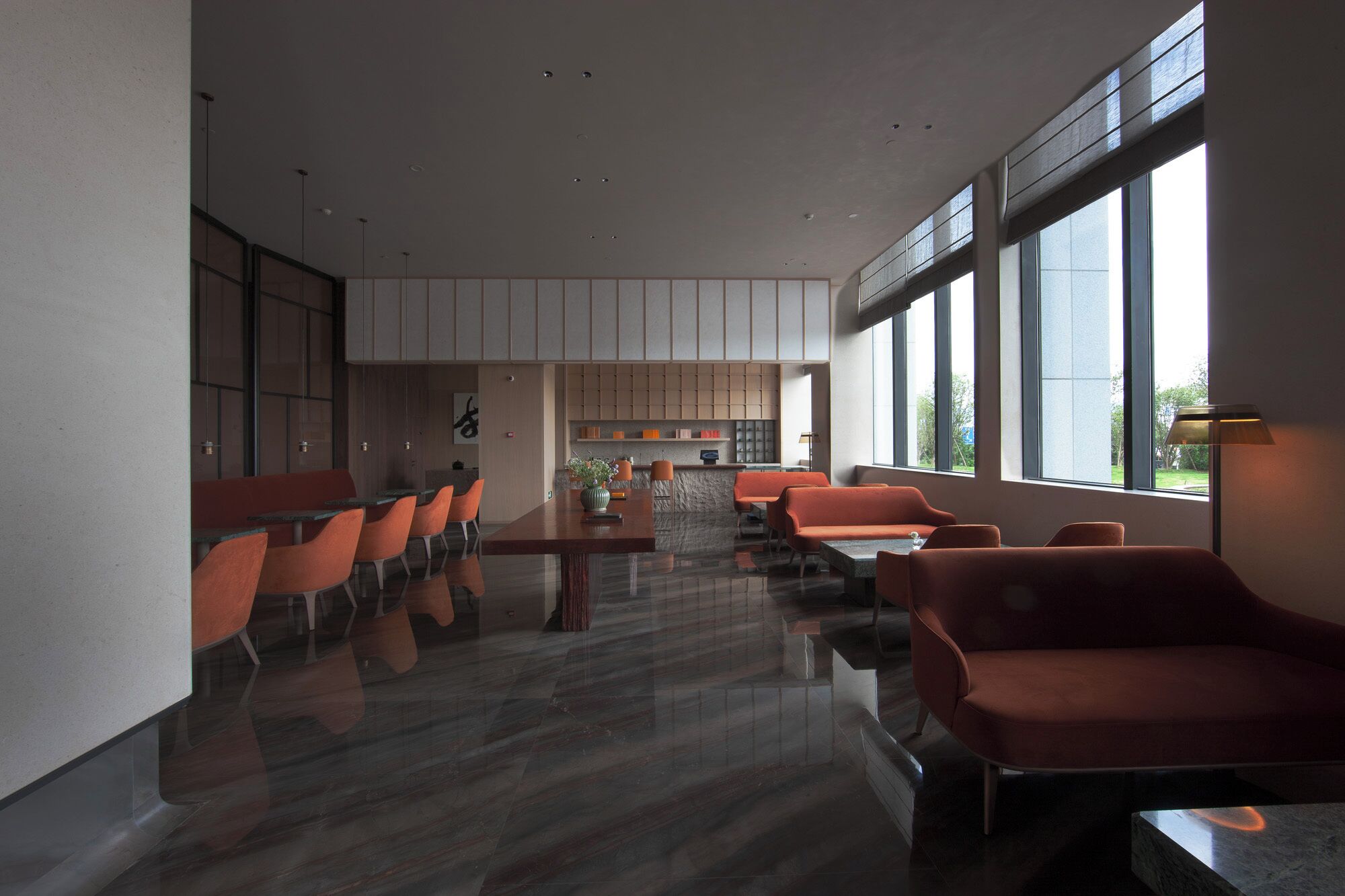 A modern dining area with a long table and orange chairs, large windows, and a kitchen in the background.