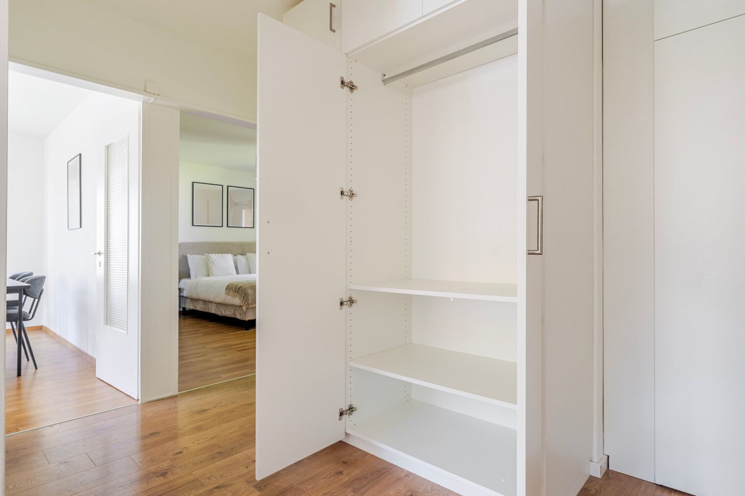 A room with a wooden floor, a bed with white bedding, and a dining area with a table and chairs.
