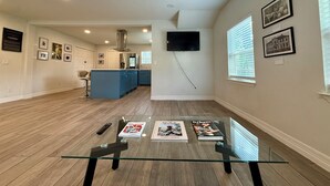 A spacious living area with a glass coffee table, a kitchen in the background, and a TV mounted on the wall. - 3rd Ward Gem: Modern Comfort + History (Houston)