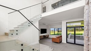 Un interior moderno con una escalera de cristal, un televisor de pantalla plana, un sofá y un ventanal con vistas a un jardín.