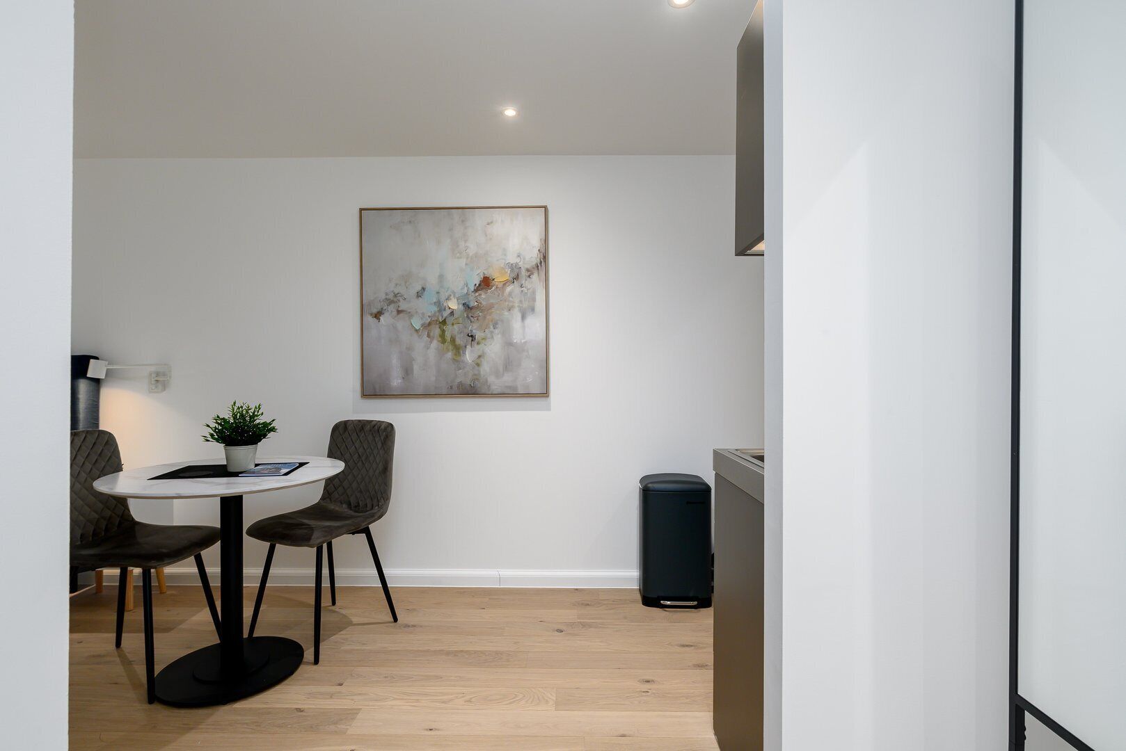 A modern dining area with a round table, two chairs, a potted plant, and a large abstract painting on the wall.